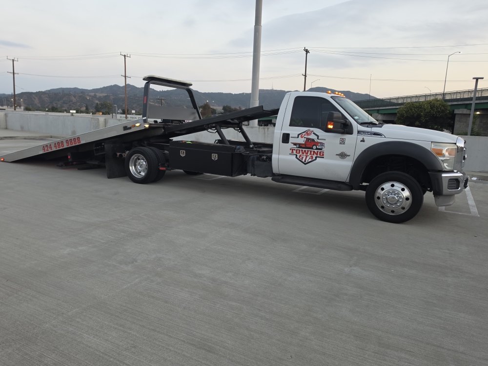 Flatbed tow truck loading a damaged car on a highway shoulder at dusk